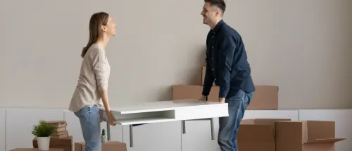 Two people lifting a white coffee table in a living room filled with moving boxes and packed belongings, illustrating the question how renters insurance covers moving.