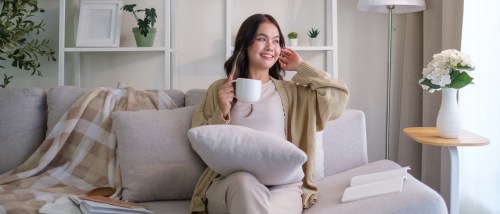 Carefree young woman sitting comfortably on a sofa, holding a mug and enjoying a peaceful moment of quiet enjoyment in her cozy home.